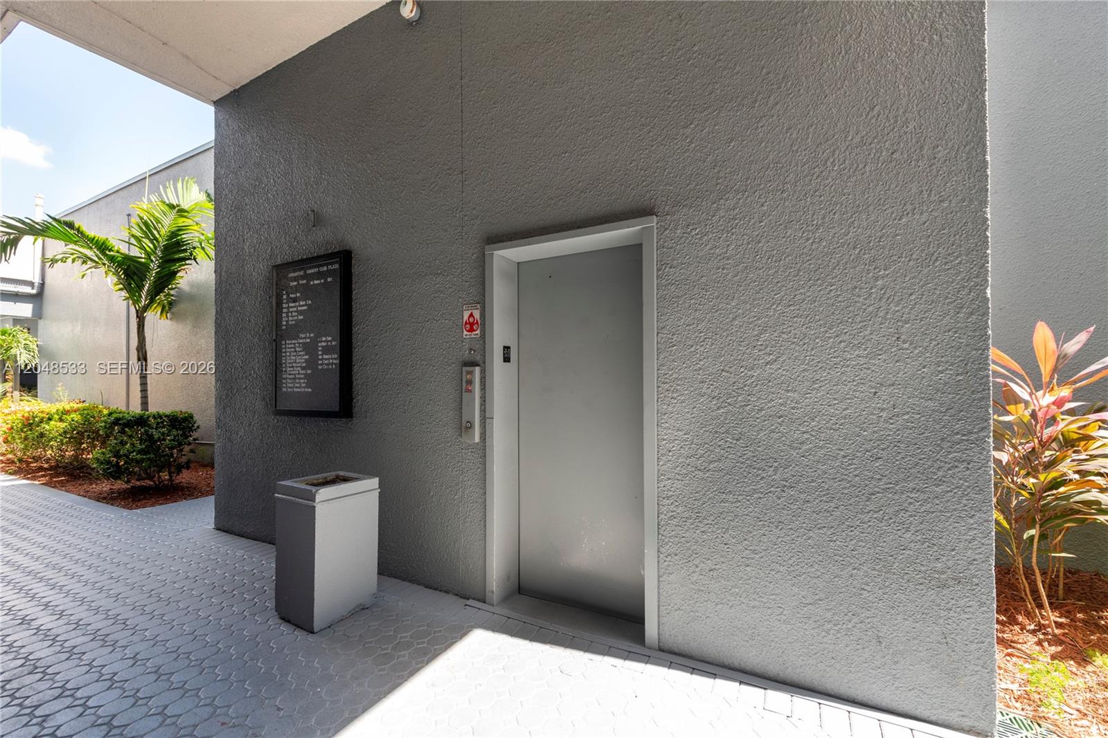 An outdoor elevator entrance features a grey stucco wall, a directory sign, and landscaping — 3801 N University Dr # 320, Sunrise FL 33351, SUNRISE, FL