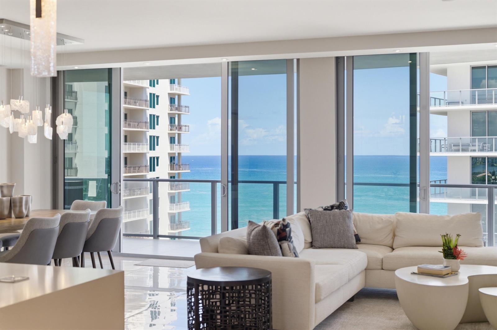 A modern living room with a white sectional sofa, dining area, and floor-to-ceiling ocean views — 3100 N Ocean Drive # 1202, Singer Island FL 33404, SINGER ISLAND, FL