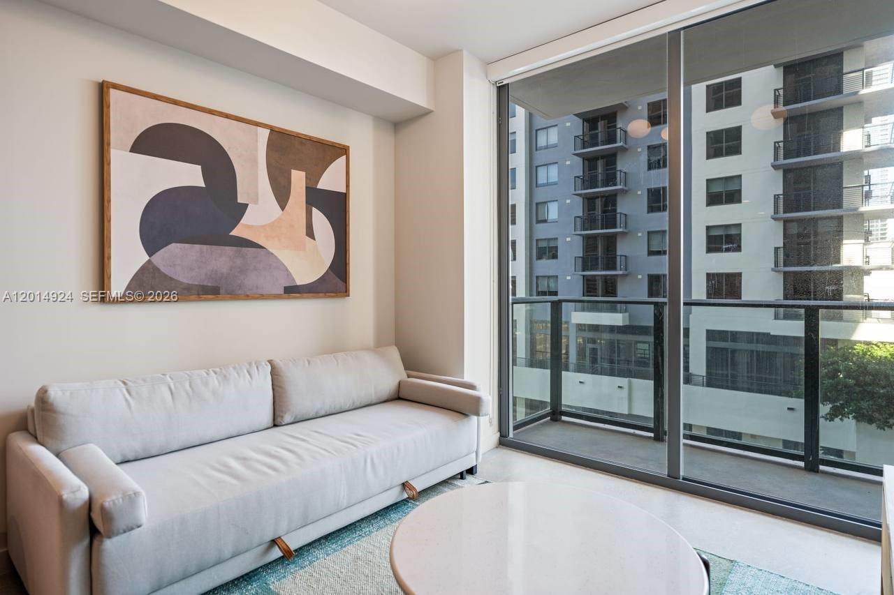 A modern living room features a light gray sofa, abstract wall art, and a balcony — 11 NE 6th # 1210, Miami FL 33132, MIAMI, FL