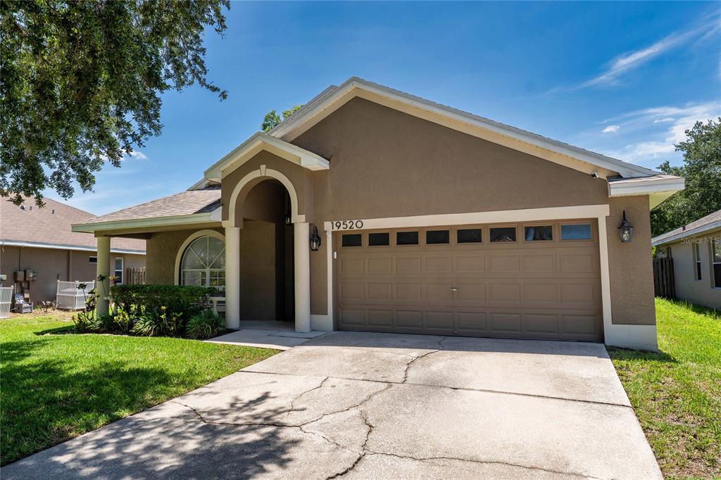 A single-story house with a tan stucco exterior, a two-car garage, and a covered entryway — 19520 MORDEN BLUSH DRIVE, LUTZ, FL