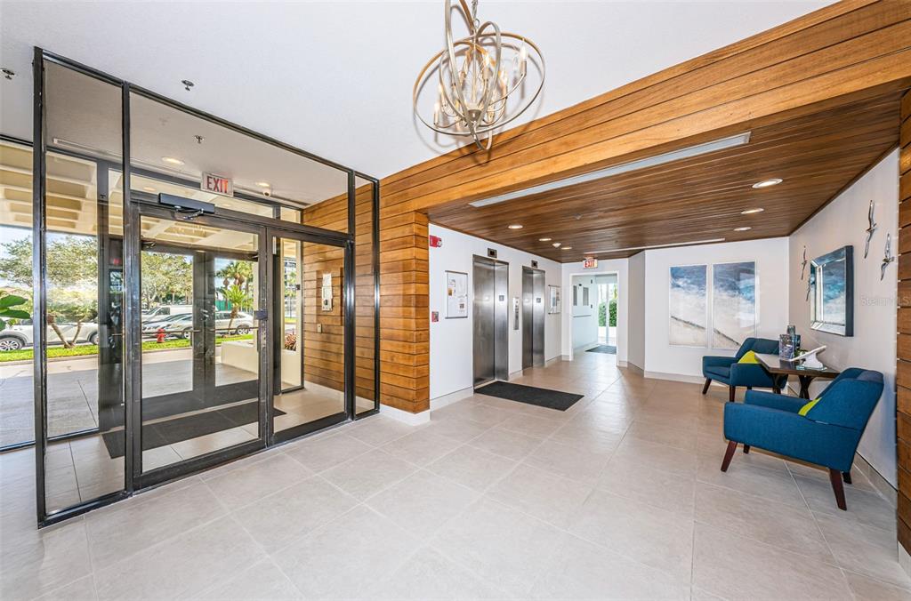 A modern lobby features a wood-paneled ceiling, two elevators, glass entry doors, and blue armchairs — 9525 BLIND PASS ROAD UNIT 505, ST PETE BEACH, FL