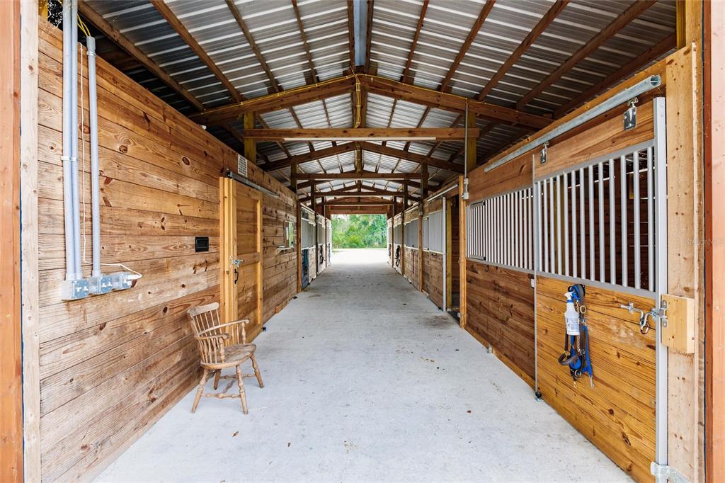 A wooden horse stable aisle features concrete flooring, multiple stalls, and a wooden chair — 9325 SW LIPE ROAD, ARCADIA, FL