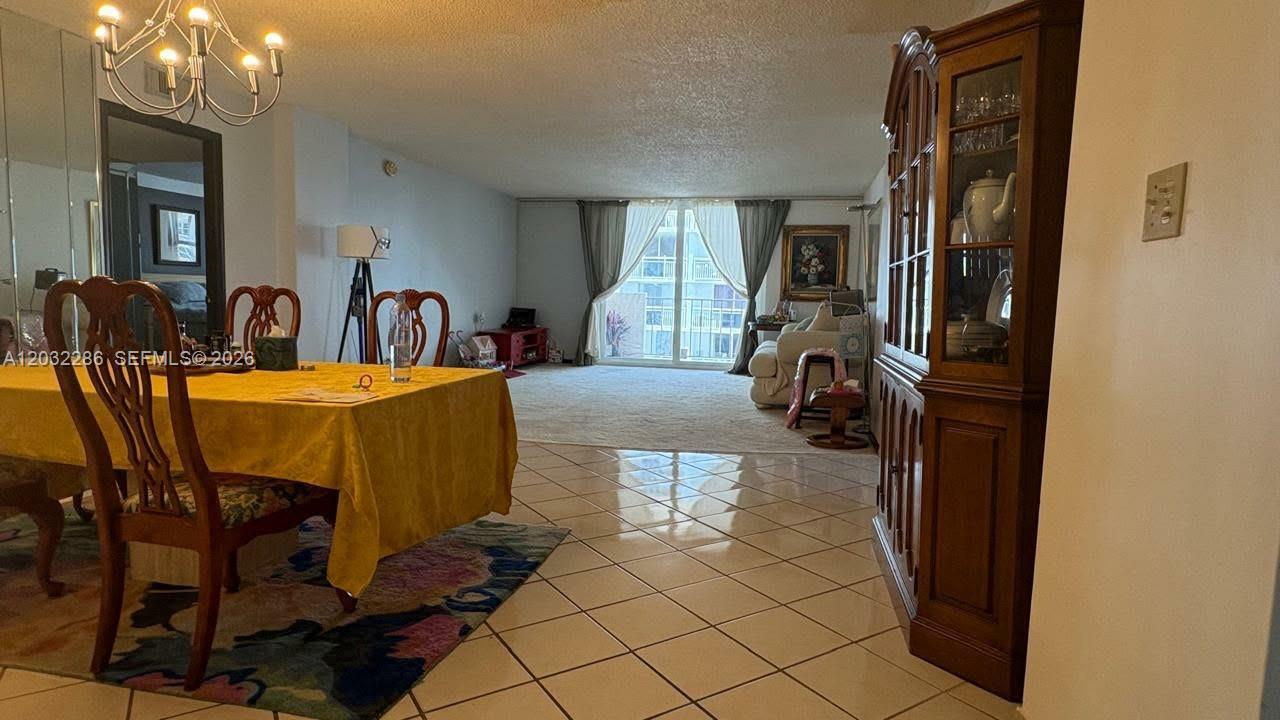 A dining area with a yellow tablecloth transitions into a carpeted living room with balcony access — 1865 S Ocean Dr # 12E, Hallandale Beach FL 33009, HALLANDALE BEACH, FL