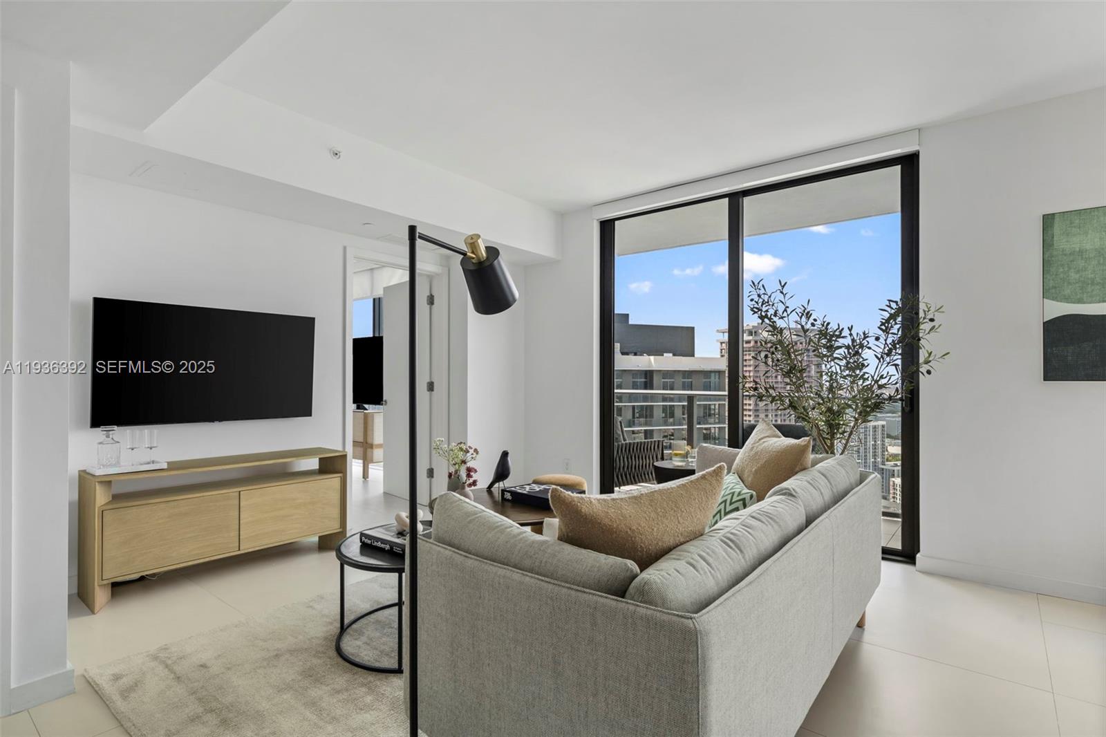 A modern living room features a grey sofa, a television, and a balcony with city views — 501 NE 31st St # 3409, Miami FL 33137, MIAMI, FL