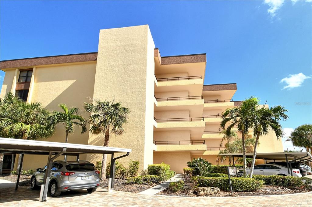 A multi-story tan apartment building features open balconies and covered parking spaces with palm trees — 8779 MIDNIGHT PASS ROAD UNIT 401H, SARASOTA, FL