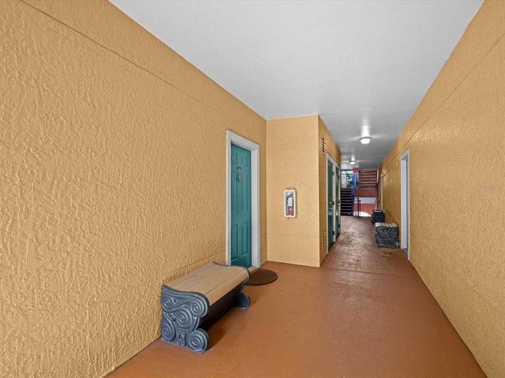An outdoor apartment hallway with yellow stucco walls, a teal door, and two decorative benches — 2705 VIA MURANO UNIT 123, CLEARWATER, FL