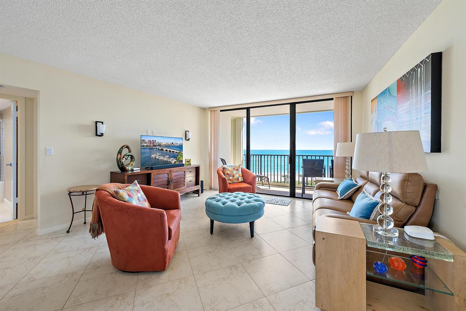 A living room features a brown leather sofa, orange armchairs, a blue ottoman, and balcony views — 400 Ocean Trail Way # 707, Jupiter FL 33477, JUPITER, FL