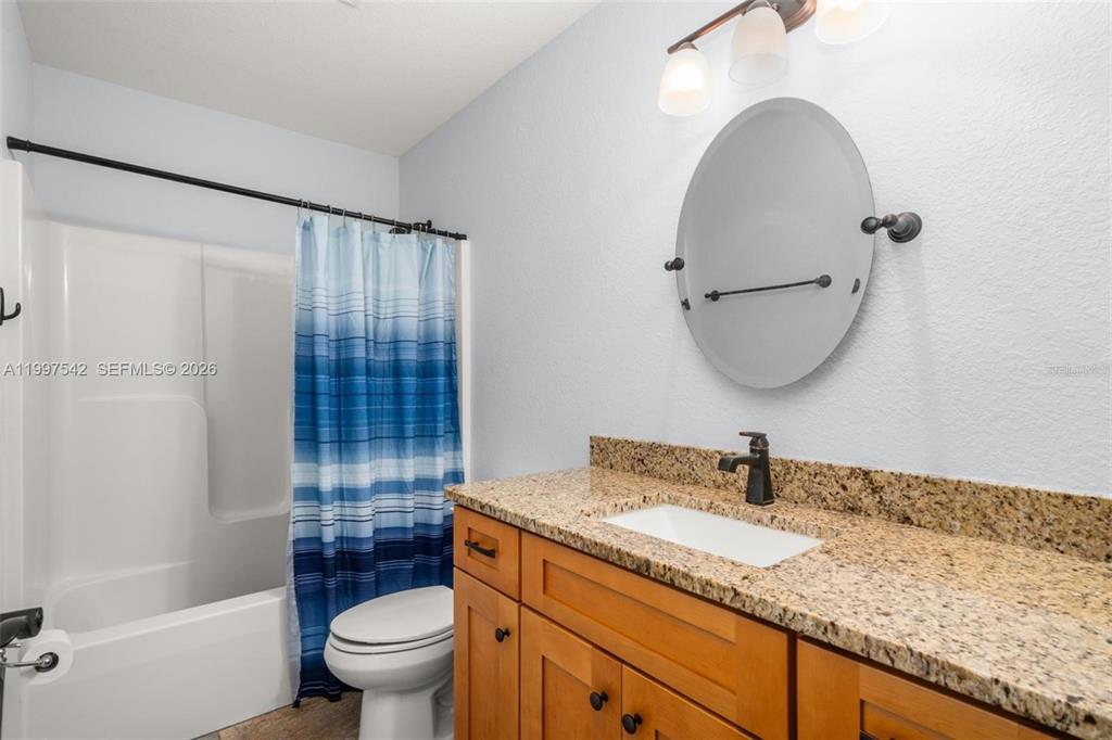 A bathroom features a granite vanity, wooden cabinets, a round mirror, and a shower-tub combination — 112 NE BIMINI STREET NE, LAKE PLACID, FL