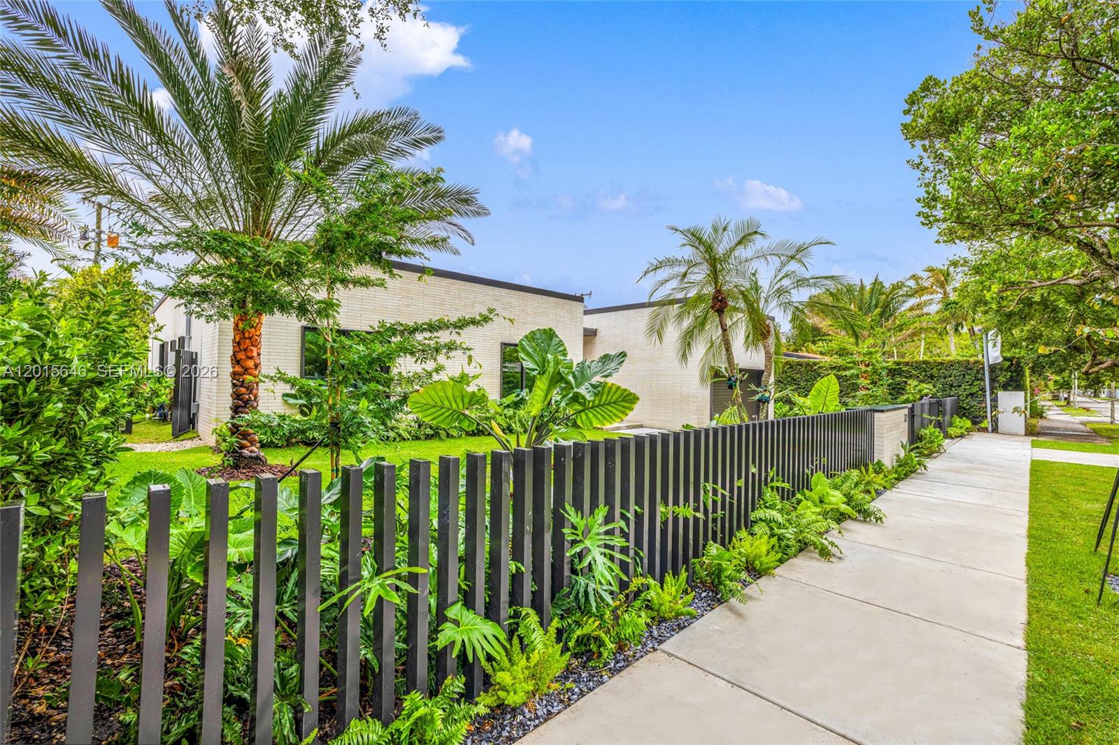 A modern, light-colored brick house is viewed from a sidewalk behind a dark picket fence — 72 W Shore Dr W, Miami FL 33133, MIAMI, FL