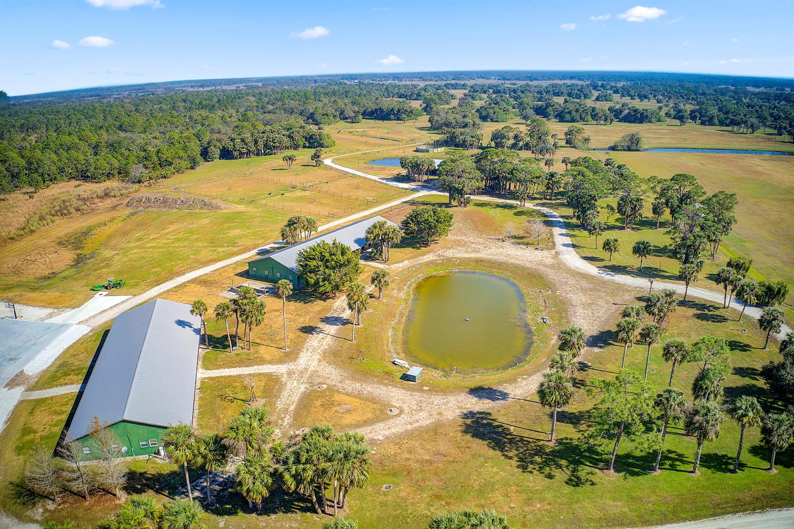 An aerial view shows two green metal barns, a pond, and palm trees on rural land — 30003 SW Martin Highway, Okeechobee FL 34974, OKEECHOBEE, FL