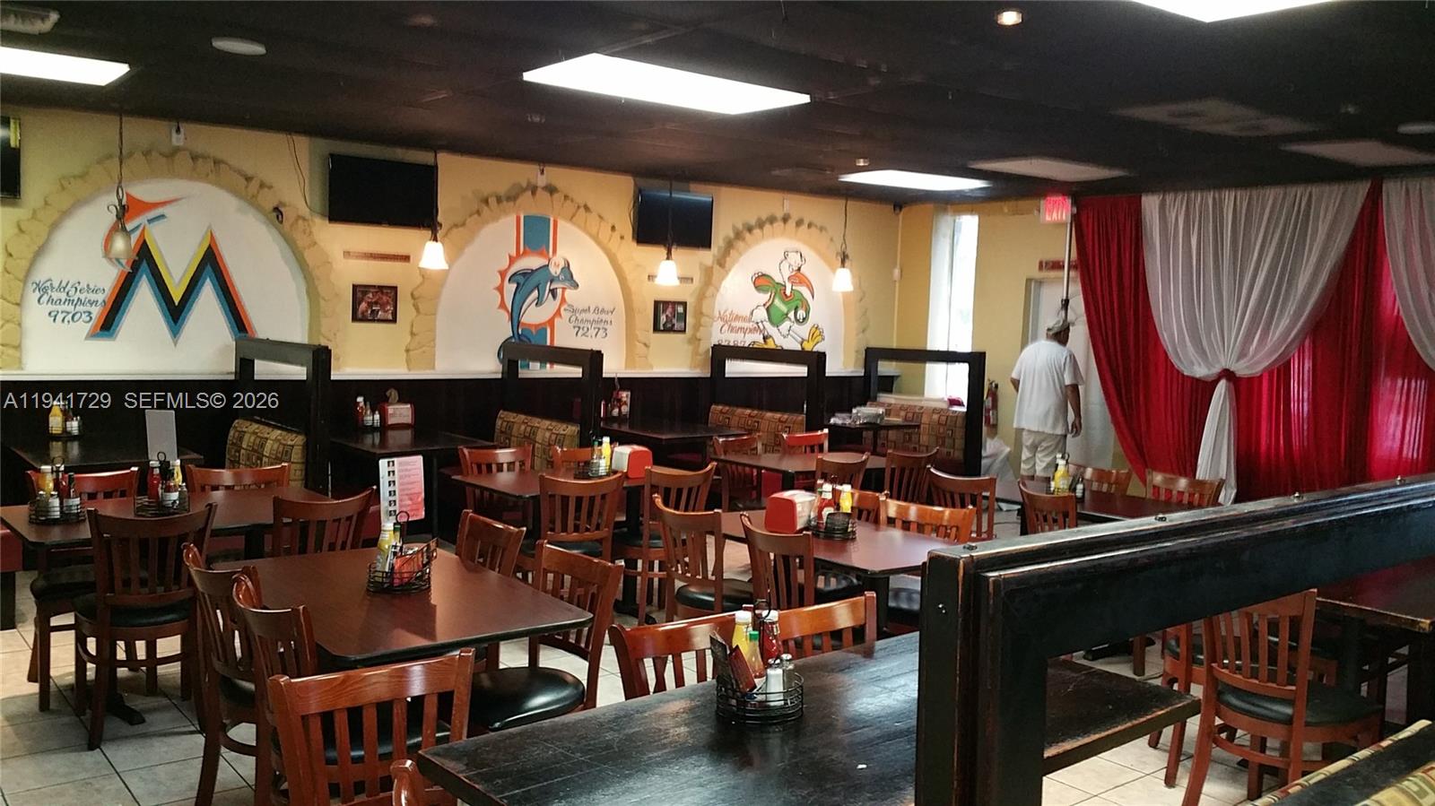 A restaurant dining room features wooden tables, chairs, and wall murals of sports team logos — 13914 NW 7th Ave, Miami FL 33168, MIAMI, FL