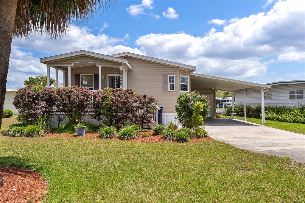 A tan mobile home with a front porch, carport, and landscaped yard under a blue sky — 2417 VINDALE ROAD, TAVARES, FL