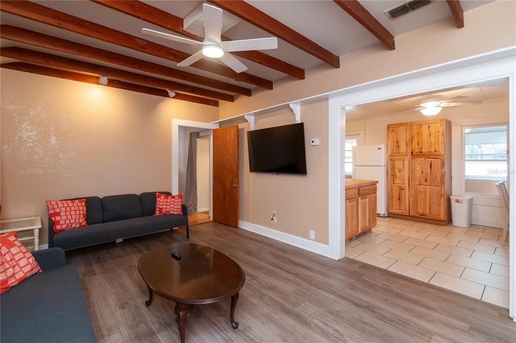 A living room with wood-look flooring, exposed ceiling beams, and an adjacent tiled kitchen area — 1235 BASS BOULEVARD, DUNEDIN, FL