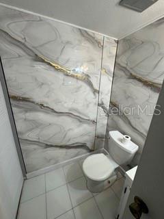 A small bathroom features a toilet and walls covered in large-format marble-patterned tile panels — 1871 PINE CONE CIRCLE UNIT 1, CLEARWATER, FL