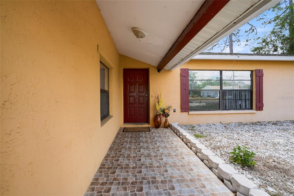 A covered porch with tiled flooring leads to a red front door and a window — 10164 GIFFORD DRIVE, SPRING HILL, FL