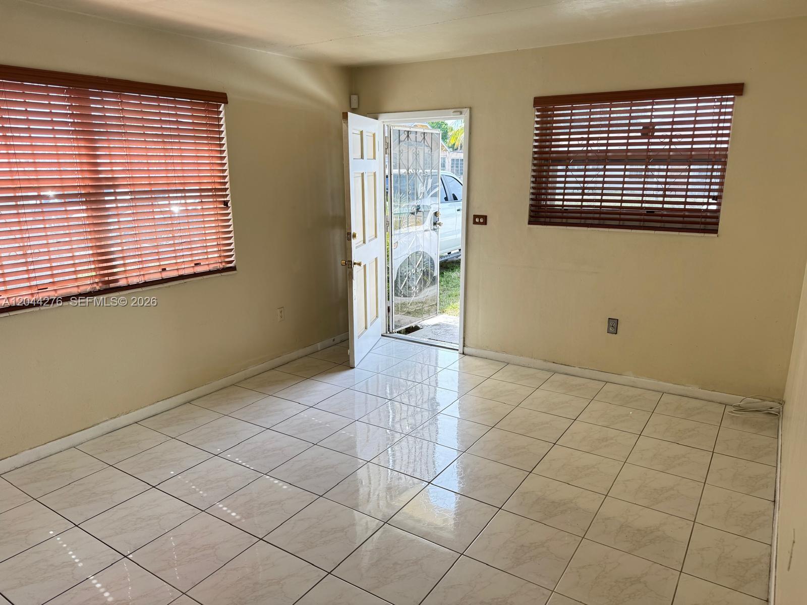 An empty room features beige walls, white tile flooring, two windows with blinds, and an open door — 2428 NW 82nd St # 2428, Miami FL 33147, MIAMI, FL