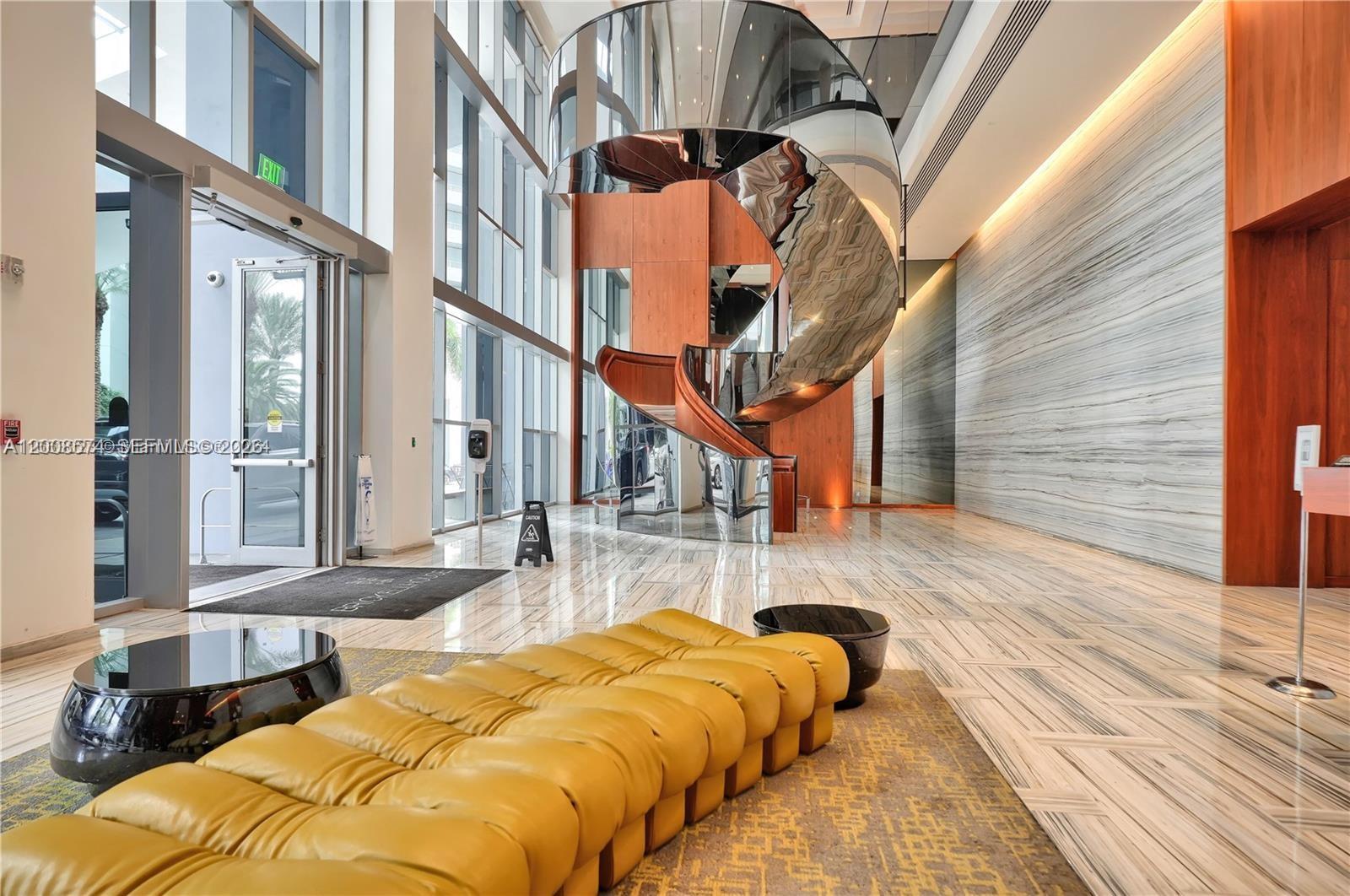 A modern lobby features a large yellow tufted bench, marble floors, and a sculptural staircase — 1300 Brickell Bay Dr # 807, Miami FL 33131, MIAMI, FL