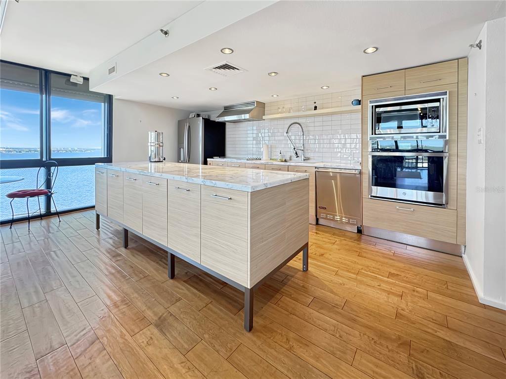 A modern kitchen features a marble-topped island, light wood cabinetry, and floor-to-ceiling windows overlooking water — 3301 BAYSHORE BOULEVARD UNIT 2003D, TAMPA, FL