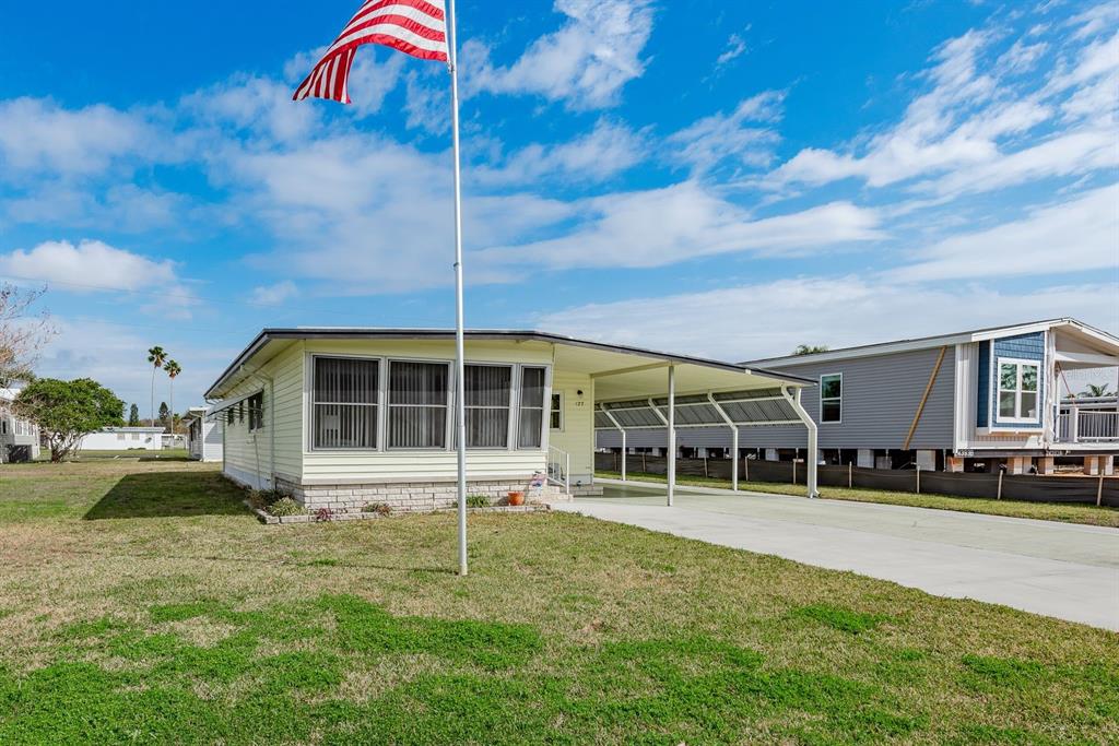 A light yellow mobile home with a carport and an American flag in the yard — 127 INDEPENDENCE AVENUE, PALM HARBOR, FL