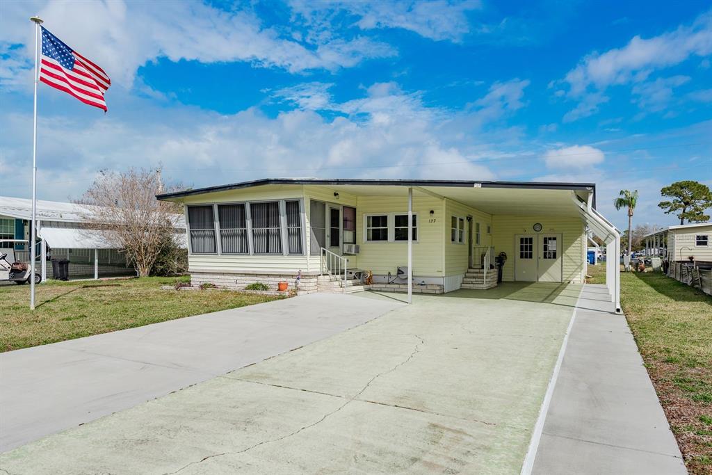 A light yellow manufactured home with a concrete driveway, carport, and an American flag — 127 INDEPENDENCE AVENUE, PALM HARBOR, FL