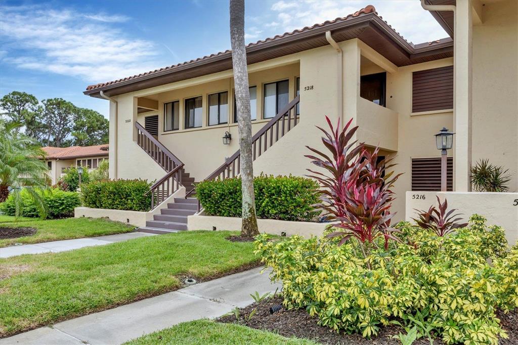 A two-story tan apartment building features an exterior staircase and lush landscaping — 5216 LAKE VILLAGE DRIVE UNIT 71, SARASOTA, FL