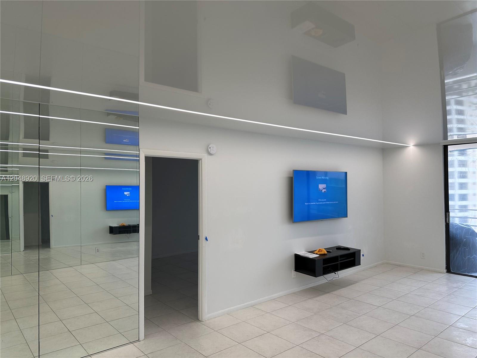 A minimalist room features white tiled floors, a mirrored wall, and a wall-mounted television — 600 Three Islands Blvd # 1113, Hallandale Beach FL 33009, HALLANDALE BEACH, FL