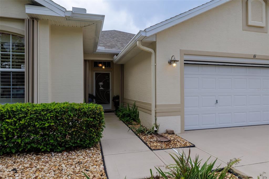 A beige house exterior features a white garage door, a front entryway, and landscaping — 61 RAINTREE CIRCLE, PALM COAST, FL
