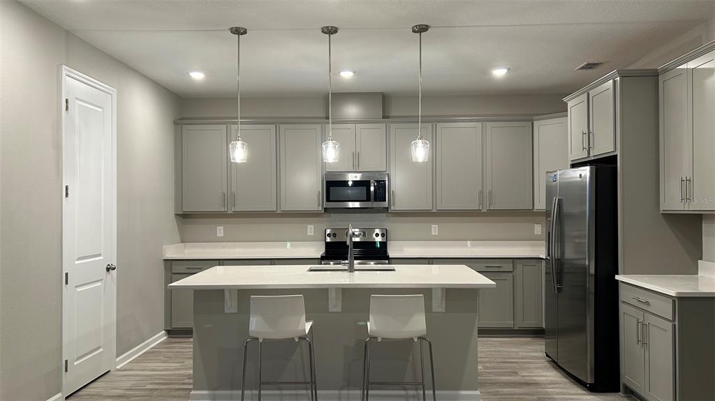 A kitchen features grey cabinets, white countertops, a central island with two stools, and stainless-steel appliances — 367 SOFIA LANE, LAKE ALFRED, FL
