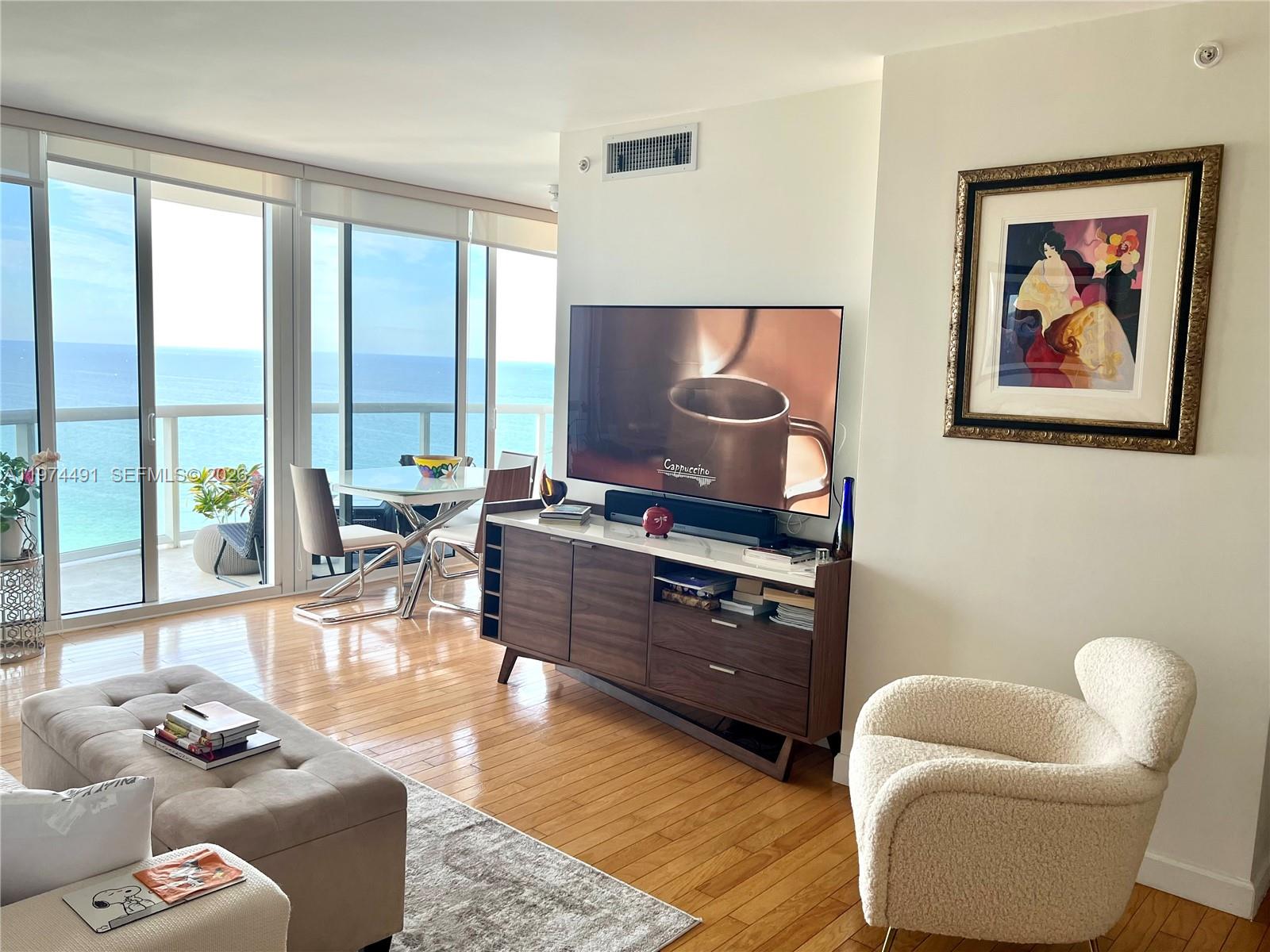 A living room features a television on a wooden console, a white armchair, and floor-to-ceiling windows — 6365 Collins Ave # 3609, Miami Beach FL 33141, MIAMI BEACH, FL