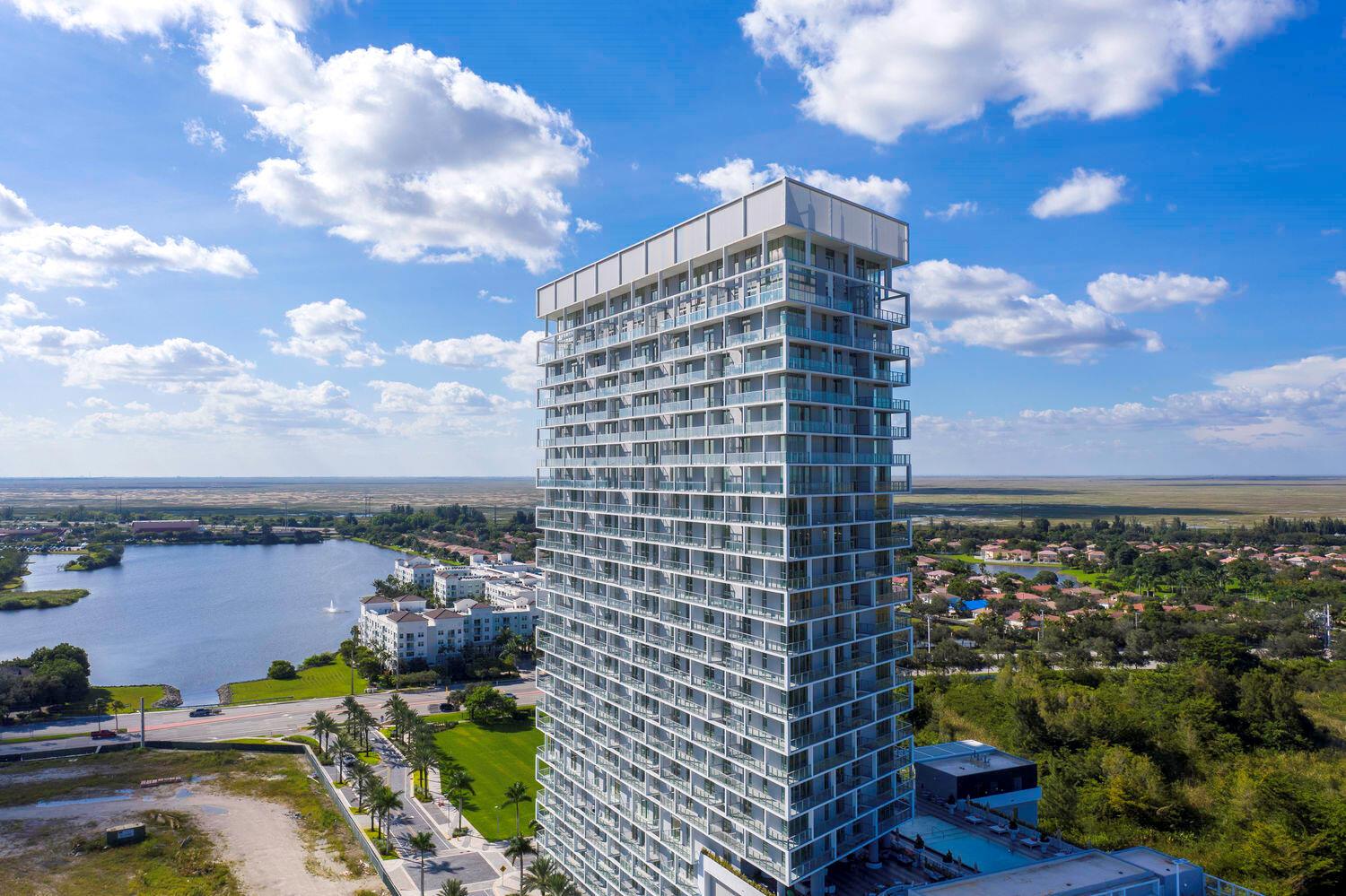 An aerial view of a tall, modern residential skyscraper surrounded by a lake and greenery — 2000 Metropica Way # 2107, Sunrise FL 33323, SUNRISE, FL