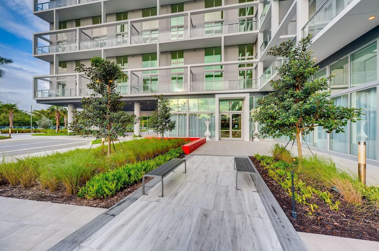 A modern apartment building exterior features a paved walkway, two benches, and landscaped garden beds — 2000 Metropica Way # 2107, Sunrise FL 33323, SUNRISE, FL