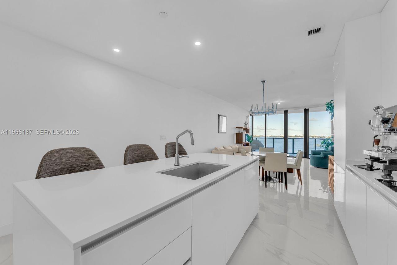 A modern kitchen with a white island and sink overlooks a living and dining area — 700 NE 24th St # 1405, Miami FL 33137, MIAMI, FL