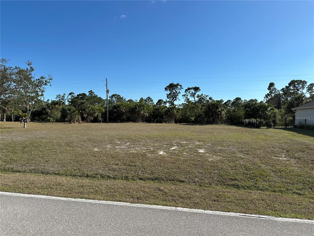 A vacant, grassy residential lot sits along a paved road under a clear blue sky — 25623 PRADA DRIVE, PUNTA GORDA, FL