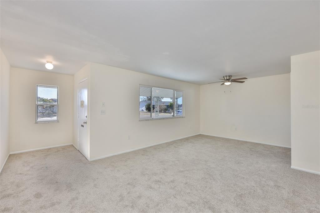 An empty living room features beige carpeting, white walls, a ceiling fan, and two windows — 703 BEL AIR AVENUE, SUN CITY CENTER, FL