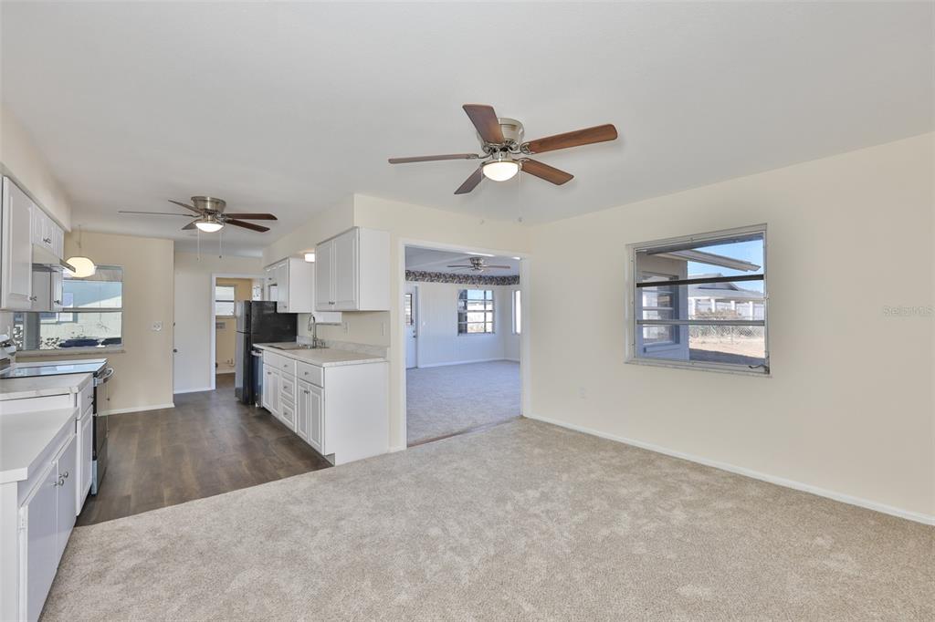 A kitchen with white cabinets and wood-look flooring opens into a carpeted living area — 703 BEL AIR AVENUE, SUN CITY CENTER, FL