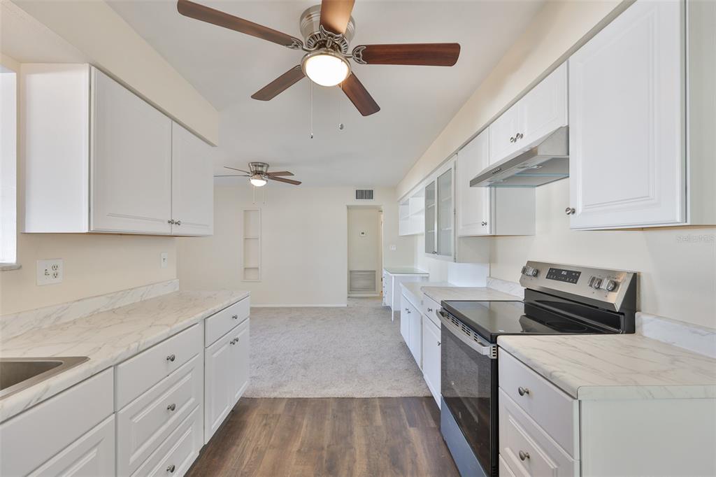 A kitchen with white cabinets, marble-look countertops, and wood-style flooring opens to a carpeted room — 703 BEL AIR AVENUE, SUN CITY CENTER, FL