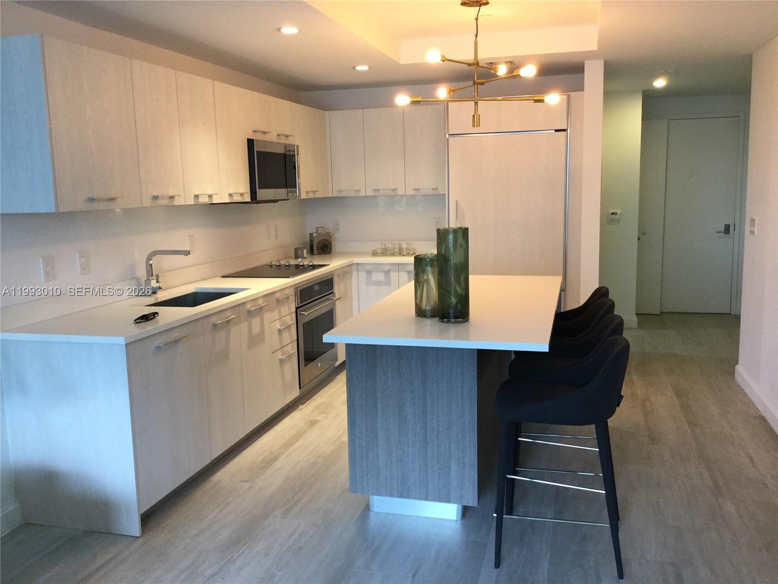 A modern kitchen features white cabinetry, a central island with bar stools, and light flooring — 55 SW 9th St # 1905, Miami FL 33130, MIAMI, FL