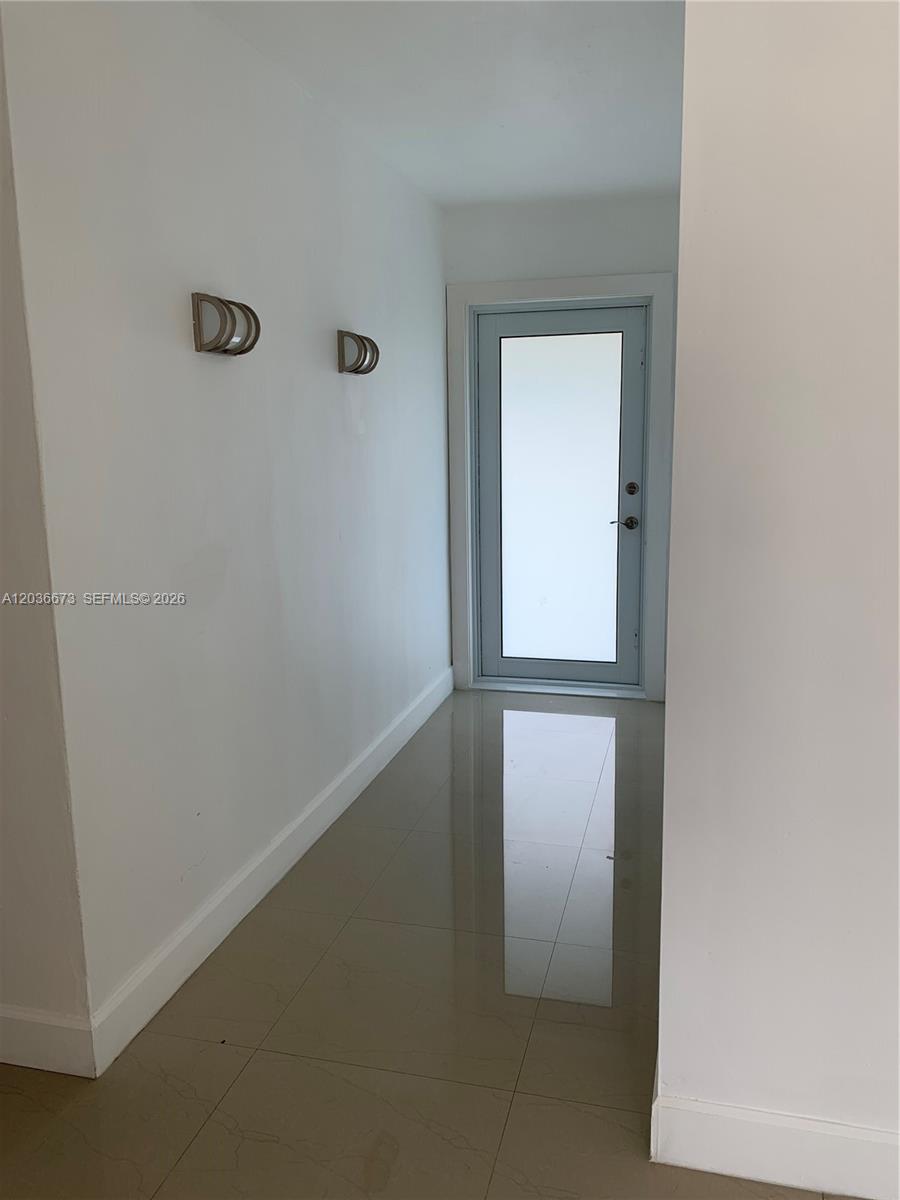 A hallway with white walls, two wall sconces, and light-colored tile flooring leads to a door — 2025 SW 84th Ct, Miami FL 33155, MIAMI, FL