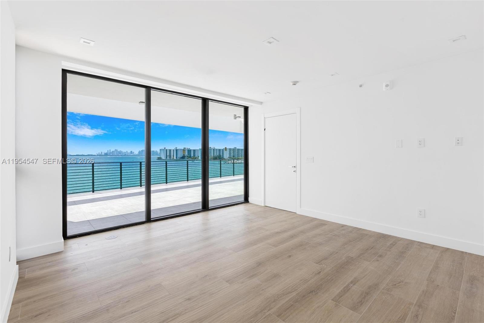 An empty room features light wood-look flooring, white walls, and floor-to-ceiling windows overlooking the water — 1930 Bay Dr Second Level Un # 1, Miami Beach FL 33141, MIAMI BEACH, FL