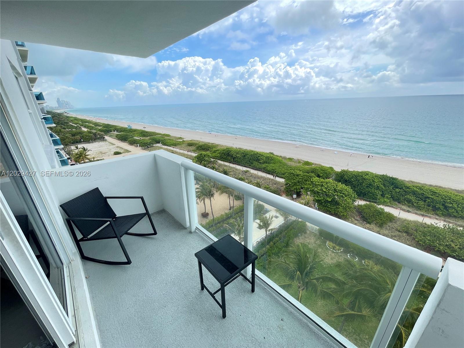 A balcony features a black rocking chair and small table overlooking the ocean and beach — 9195 Collins Ave # 1001, Surfside FL 33154, SURFSIDE, FL