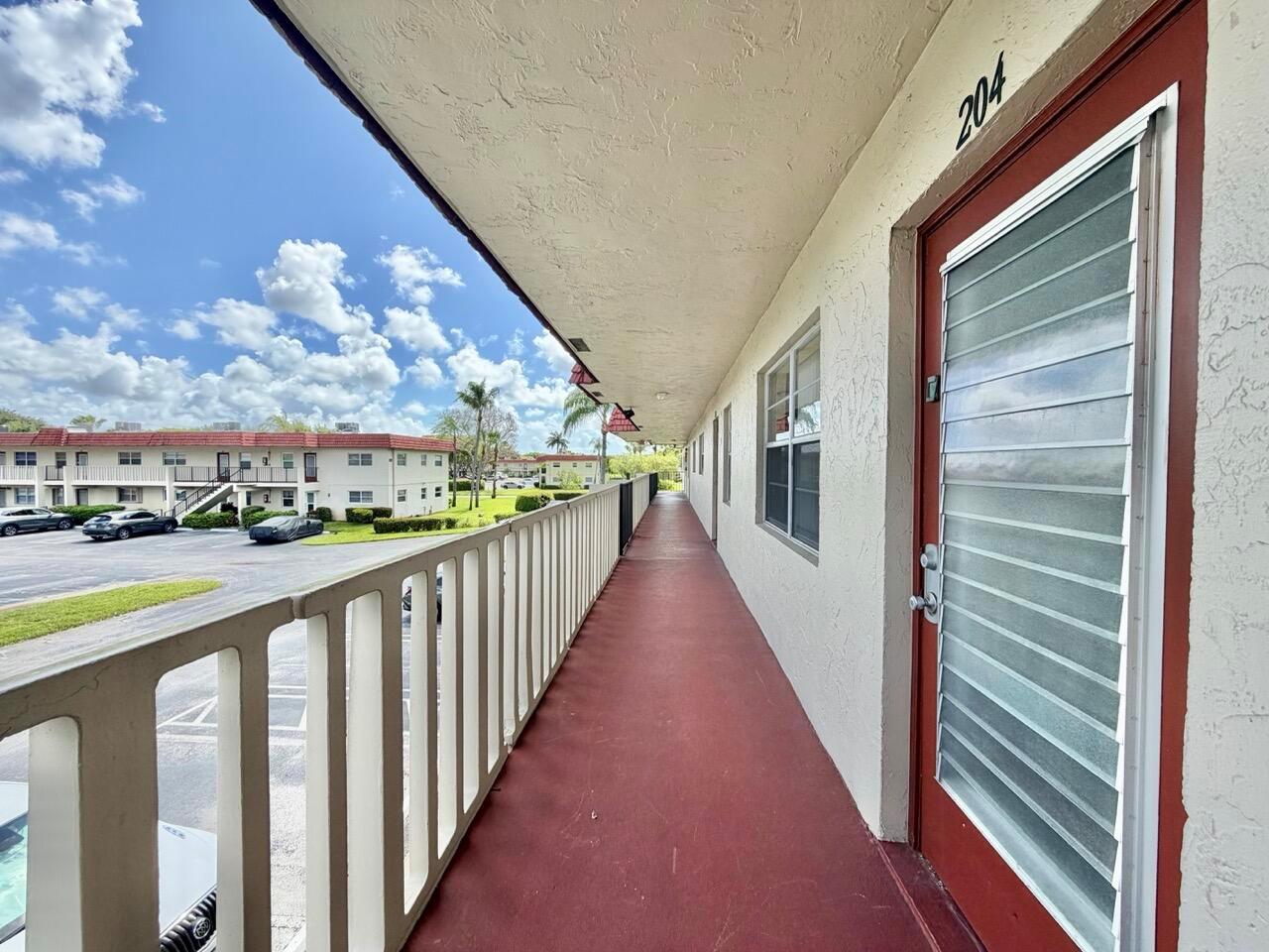 An exterior walkway with a red floor and white railing leads to a door numbered 204 — 19 Abbey Lane # 204, Delray Beach FL 33446, DELRAY BEACH, FL