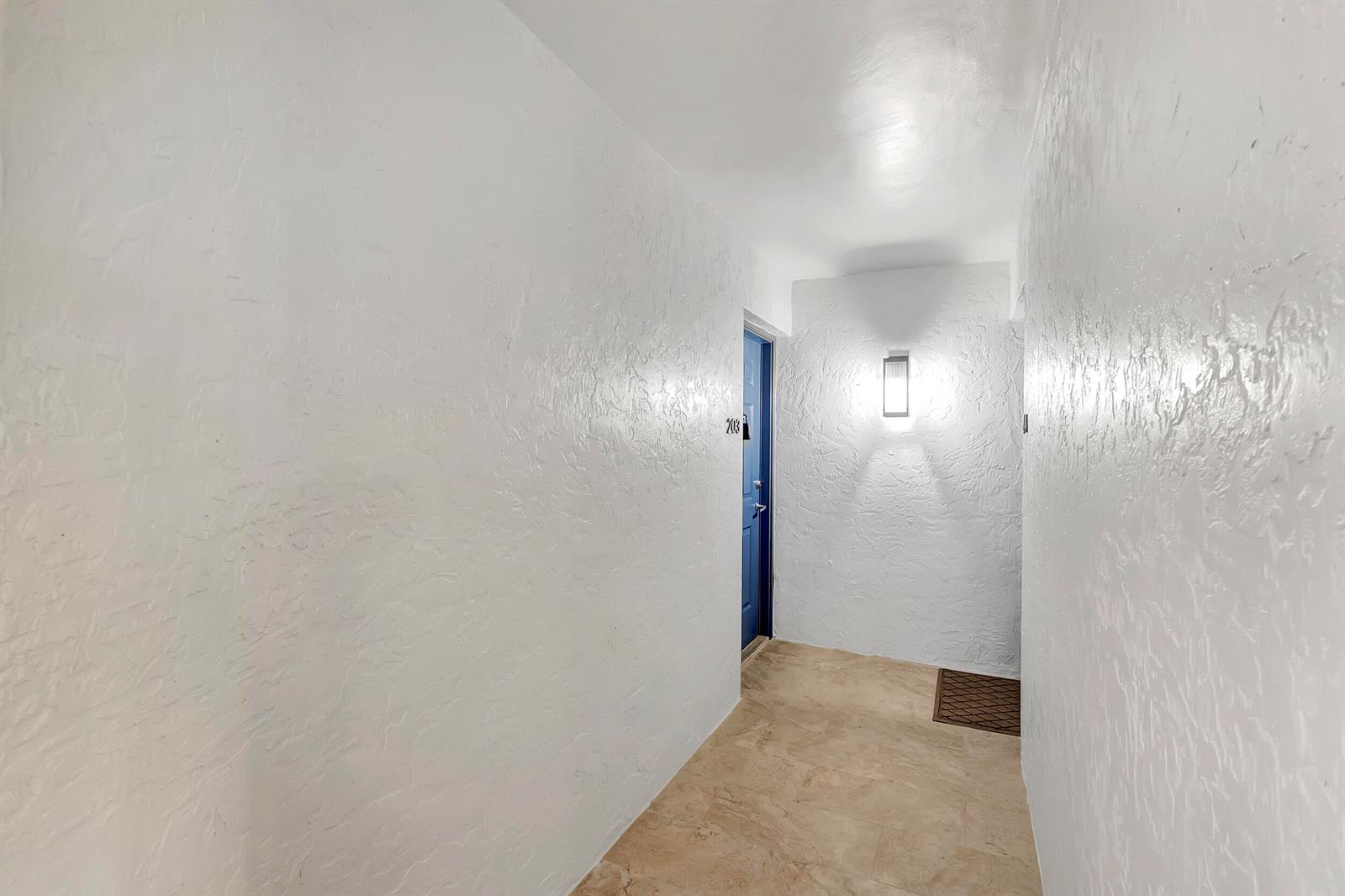 A narrow hallway with white textured walls, beige tile flooring, and a blue apartment door — 3111 Clint Moore Road # 203, Boca Raton FL 33496, BOCA RATON, FL