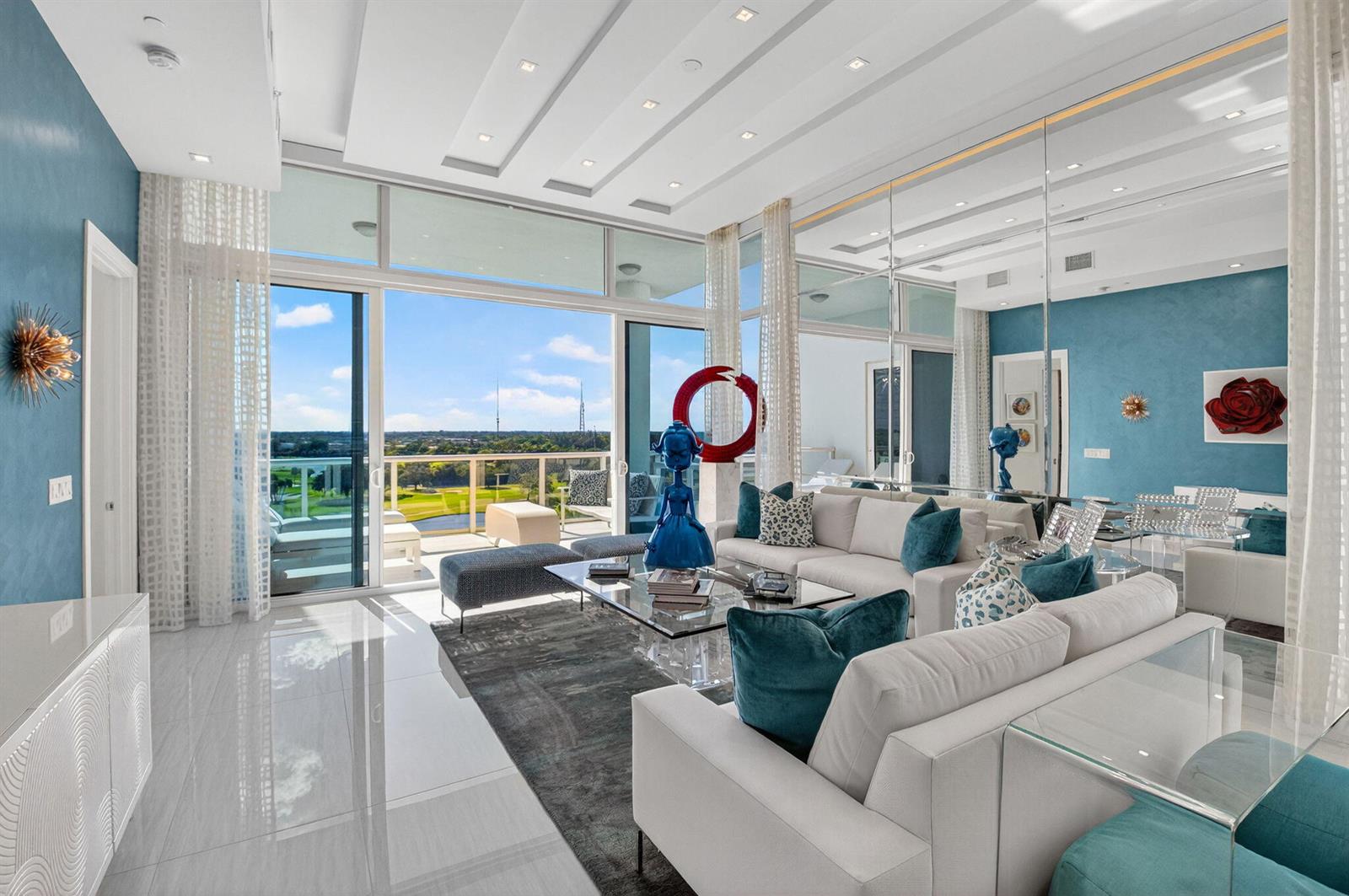 A modern living room features white sofas, blue accent walls, mirrored panels, and a balcony view — 20155 Boca West Drive # b904, Boca Raton FL 33434, BOCA RATON, FL