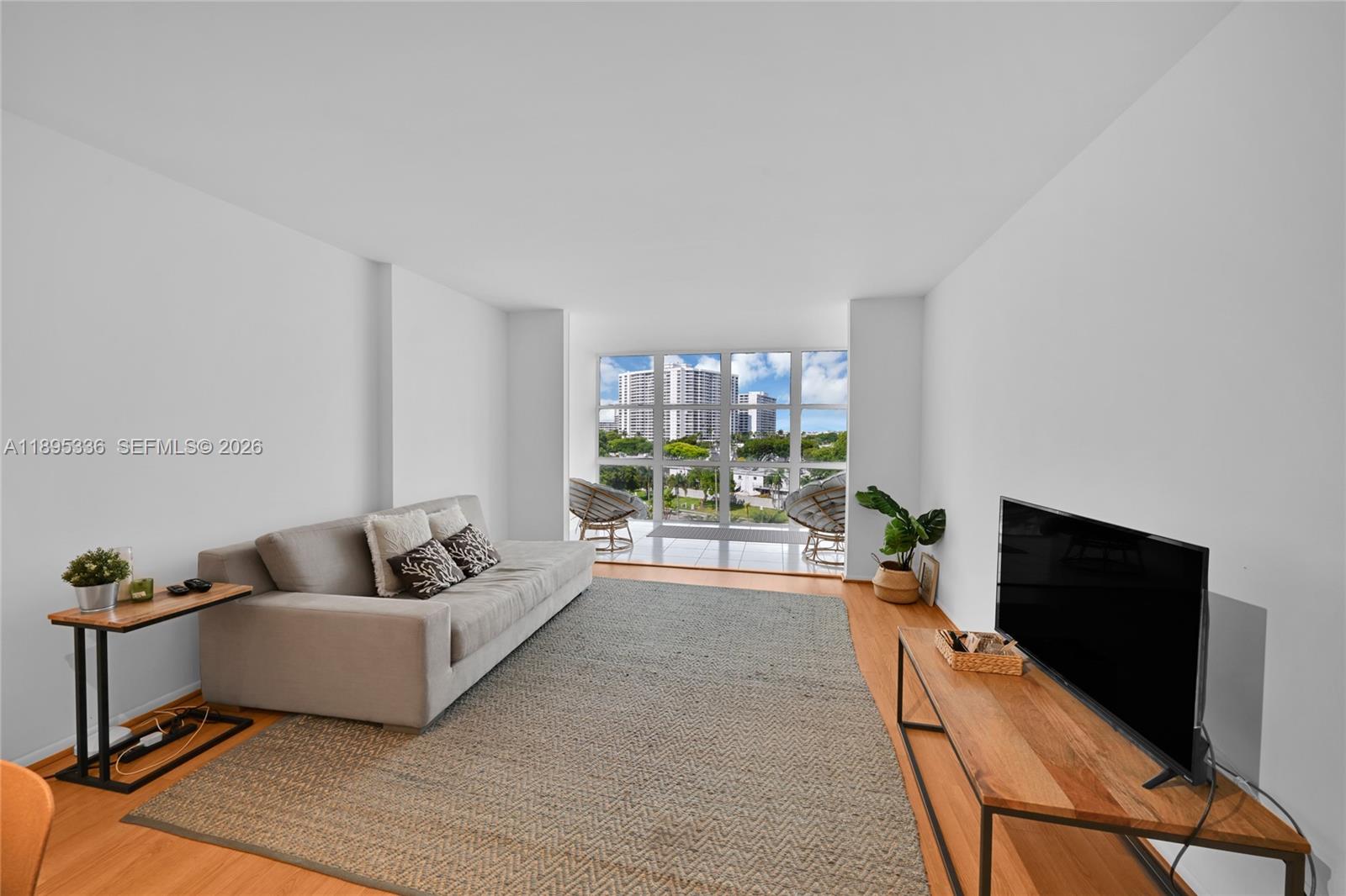 A living room features a grey sofa, a wooden TV stand, and large floor-to-ceiling windows — 800 Parkview Dr # 605, Hallandale Beach FL 33009, HALLANDALE BEACH, FL