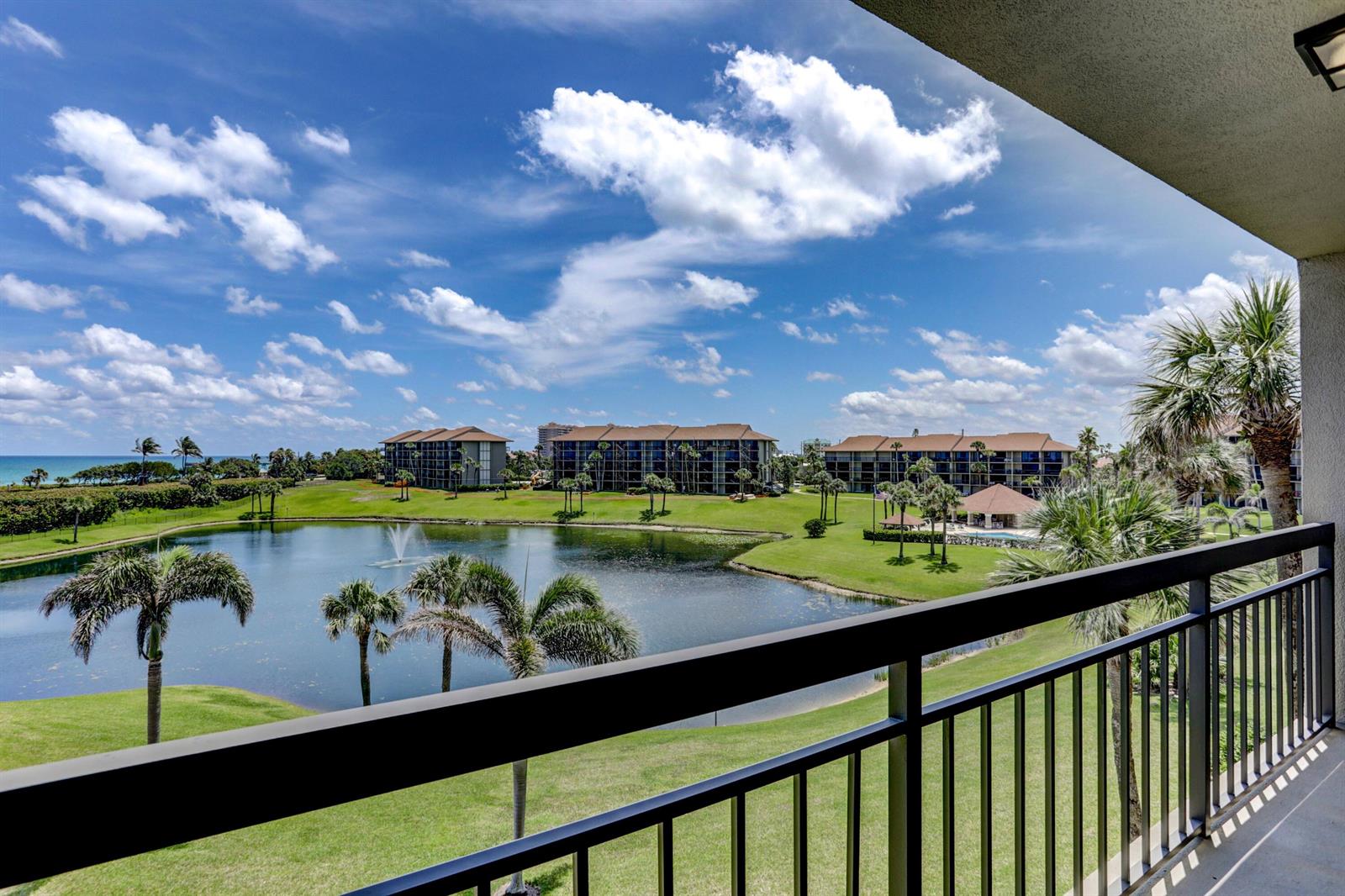 A balcony view overlooking a pond, palm trees, and multi-story residential buildings under a blue sky — 201 S Seas Drive # 401, Jupiter FL 33477, JUPITER, FL