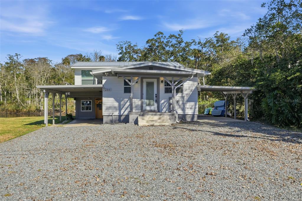 A light-colored house with a metal roof and two carports sits on a gravel lot — 7266 TROPICAL DRIVE, WEEKI WACHEE, FL