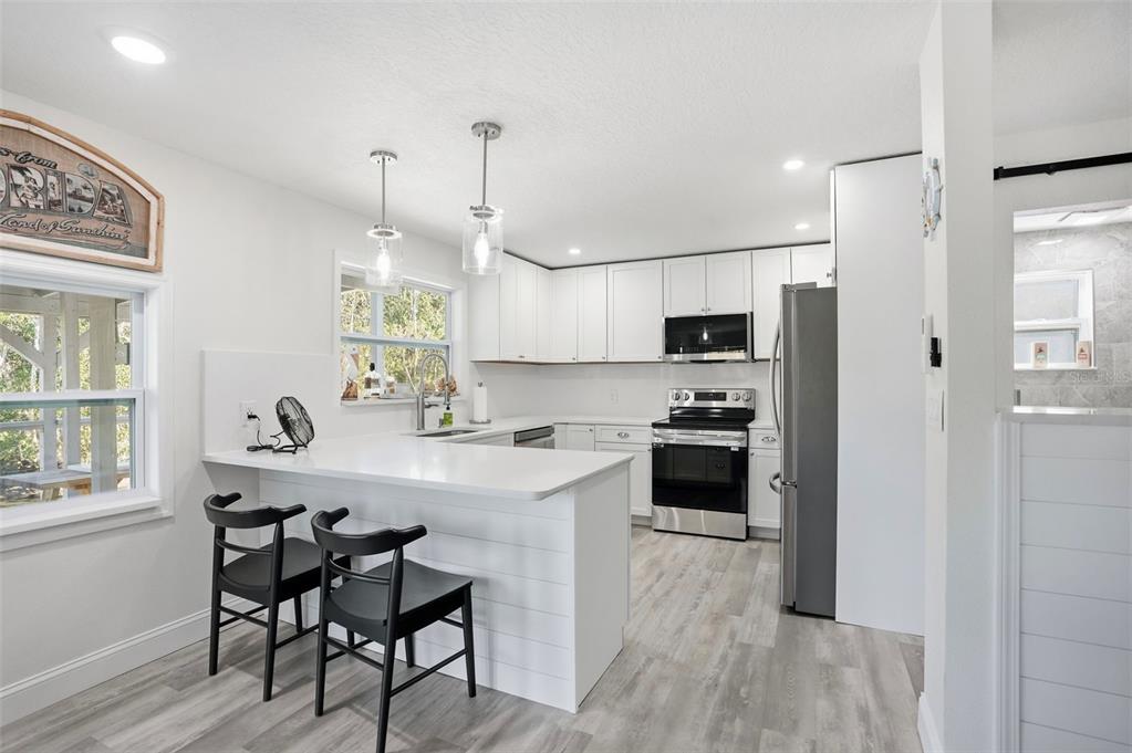 A white kitchen features a peninsula with bar stools, stainless appliances, and light wood flooring — 7266 TROPICAL DRIVE, WEEKI WACHEE, FL