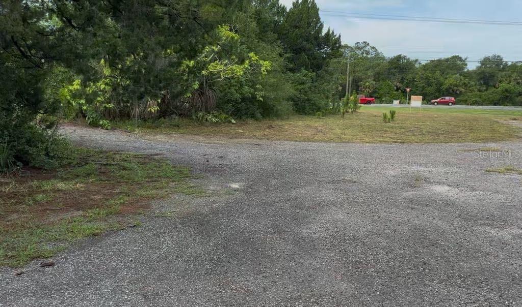 A gravel driveway leads toward a grassy area bordered by trees and a road — 3493 S RIDGEWOOD AVENUE, EDGEWATER, FL