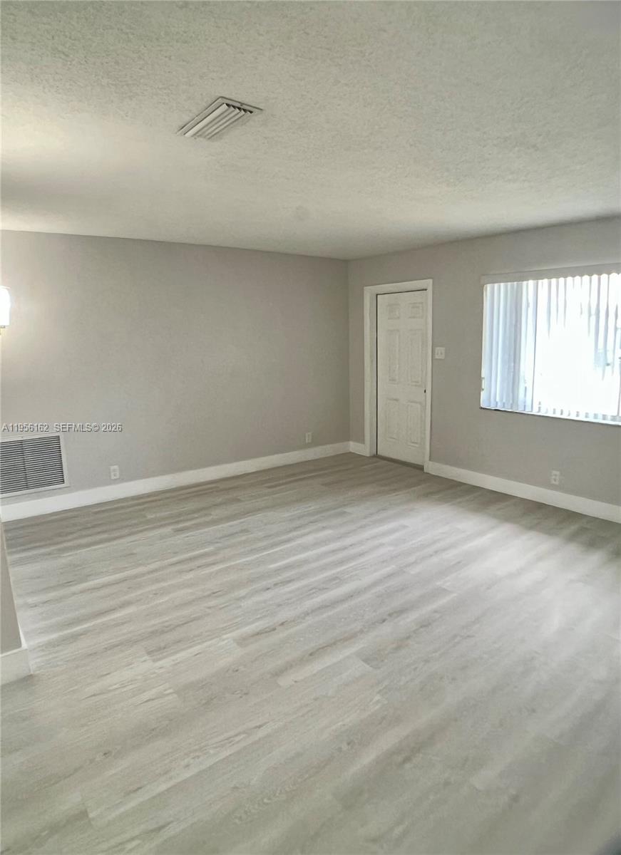 An empty room features light-colored wood-look flooring, gray walls, a white door, and vertical blinds — 6990 NW 17th St, Margate FL 33063, MARGATE, FL