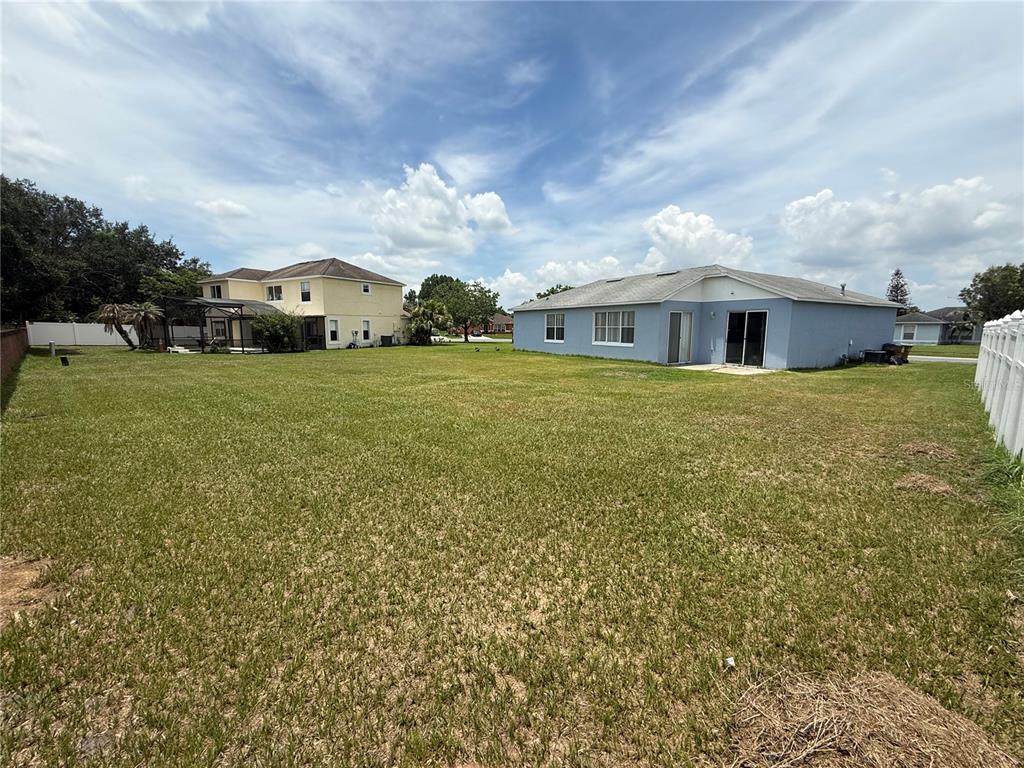 A large, grassy backyard sits between two residential houses under a blue, cloudy sky — 2560 SAGE DRIVE W, KISSIMMEE, FL